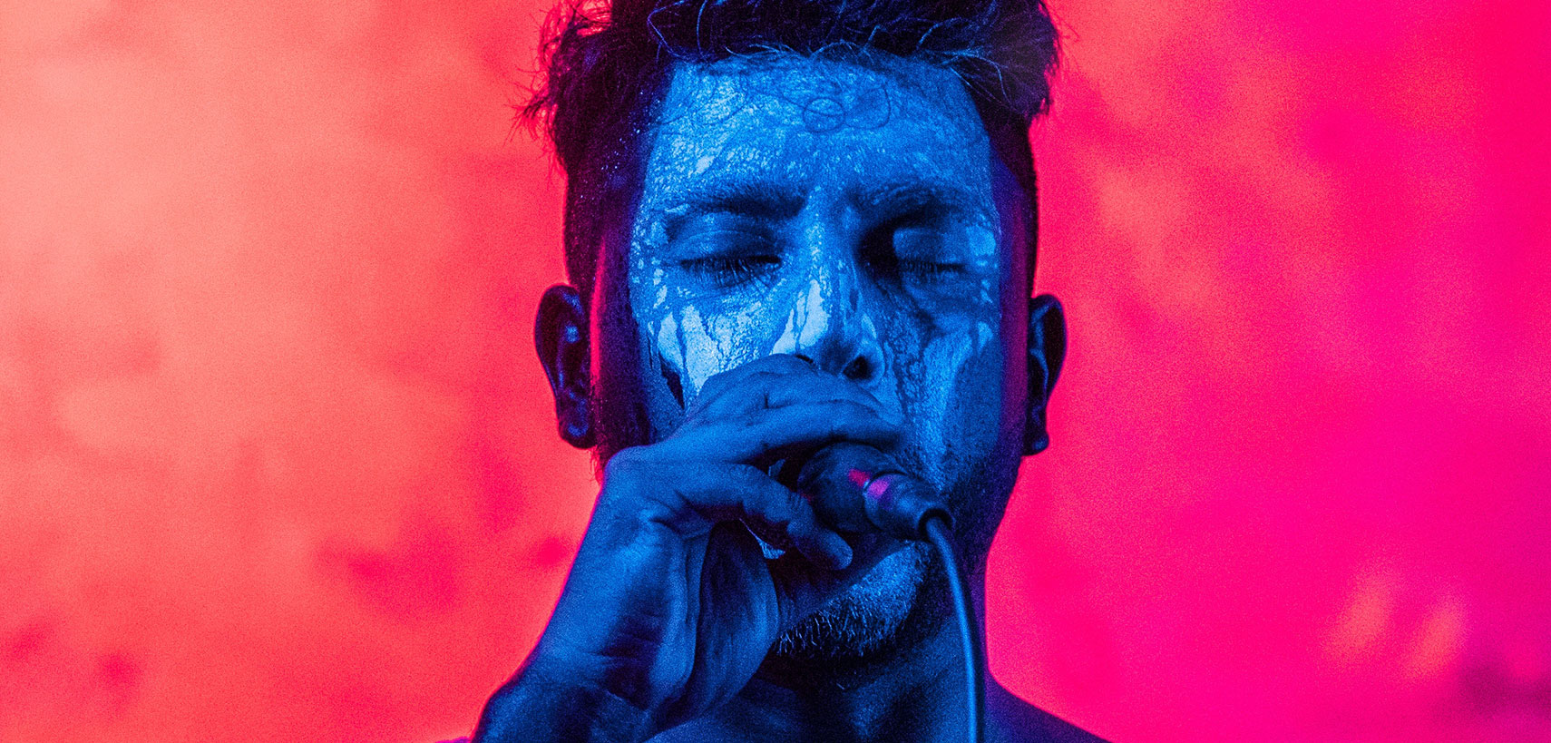 Portrait of a man with dramatic blue and red lighting, eyes closed and hand near mouth.