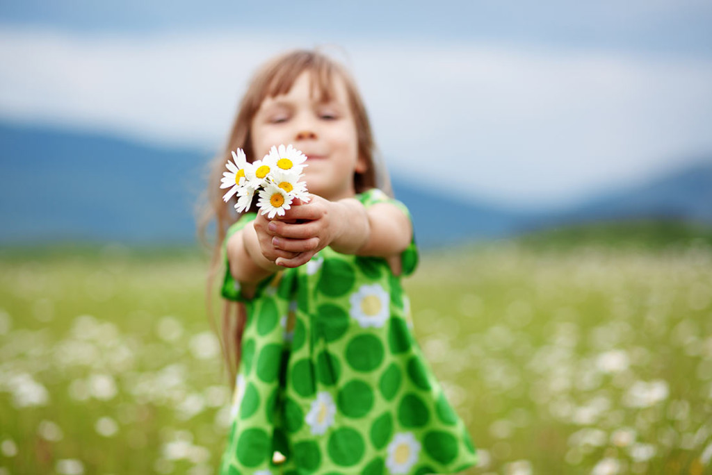 A young girl in a green dress holding out daisies.