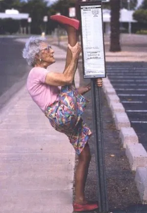 Elderly woman stretching by holding a pole on the sidewalk.